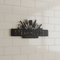 Personalized steakhouse metal sign with crossed knives, cleaver and name in black steel